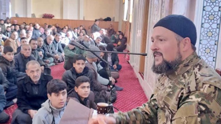 A preacher appears wearing a military uniform and a gun on the pulpit in Nawa
